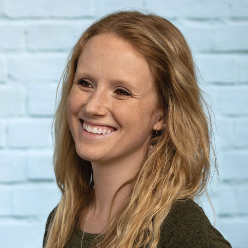 Person with long blond hair smiling, wearing a dark green top, standing against a light blue brick wall.